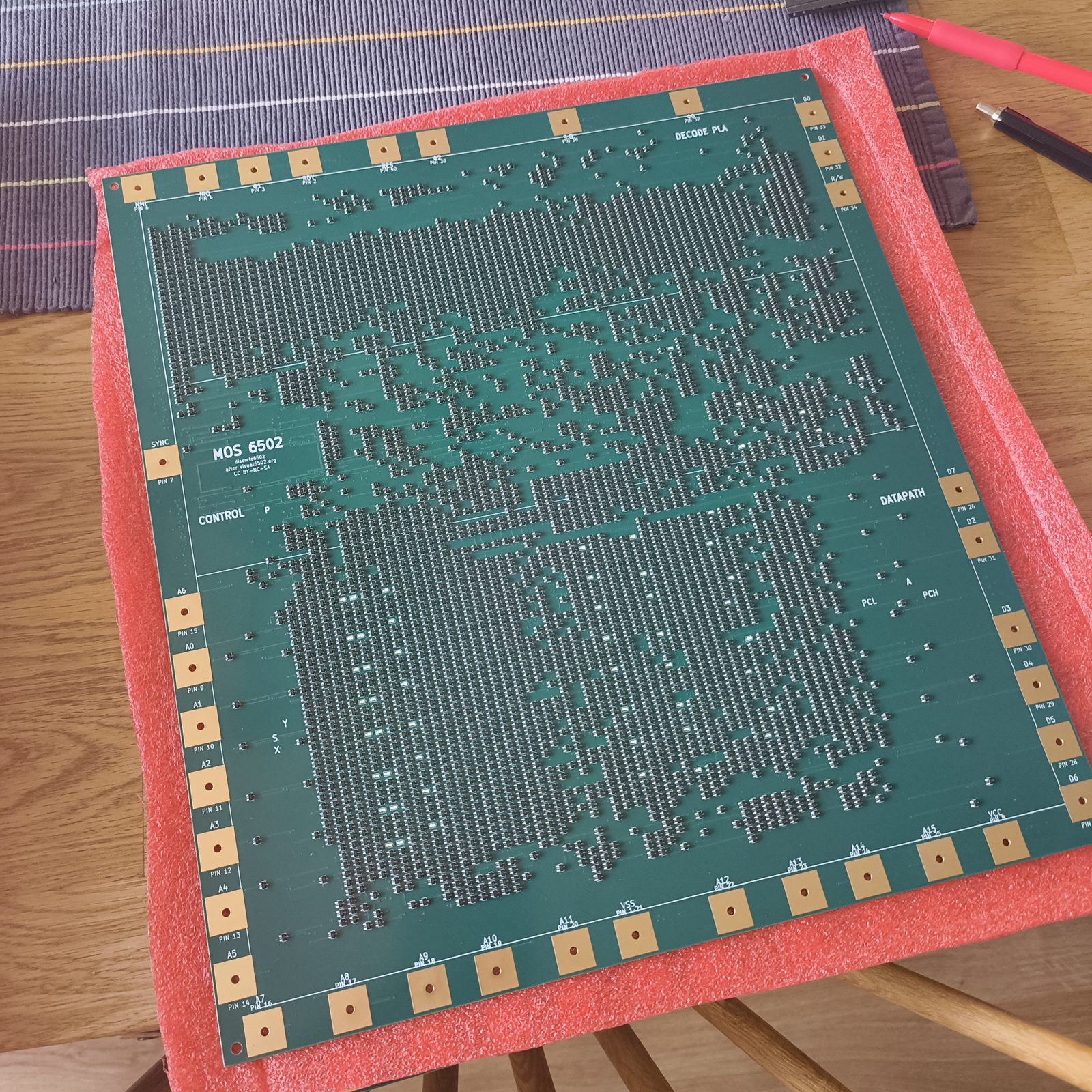 The assembled discrete6502 board on pink antistatic foam on a wooden table, a pen
                  and pencil beside it for scale. Thousands of tiny black surface-mount transistors
                  cover the green board in the pattern of the 6502 die, with gold bond pads around
                  the edge.