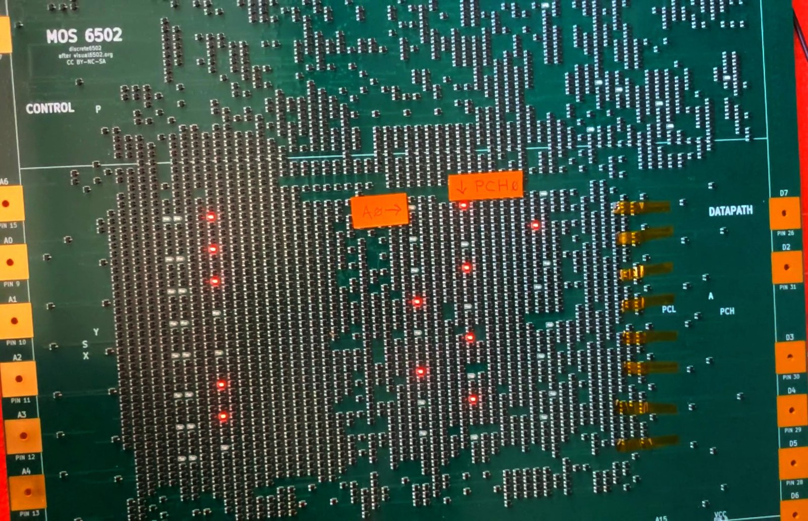 Close-up of the powered board. Red LEDs are lit in a vertical column among the
                  transistor array. Two hand-written orange sticky labels point at individual LEDs,
                  marked A0 and PCH0. A column of amber Kapton tape patches runs down the right of
                  the array where eight transistors have been reworked by hand.